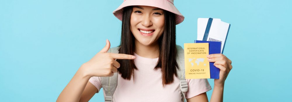 Vaccinated asian tourist, girl showing international health passport, tickets for tour abroad, smiling happy, travelling during covid-19 pandemic, blue background.