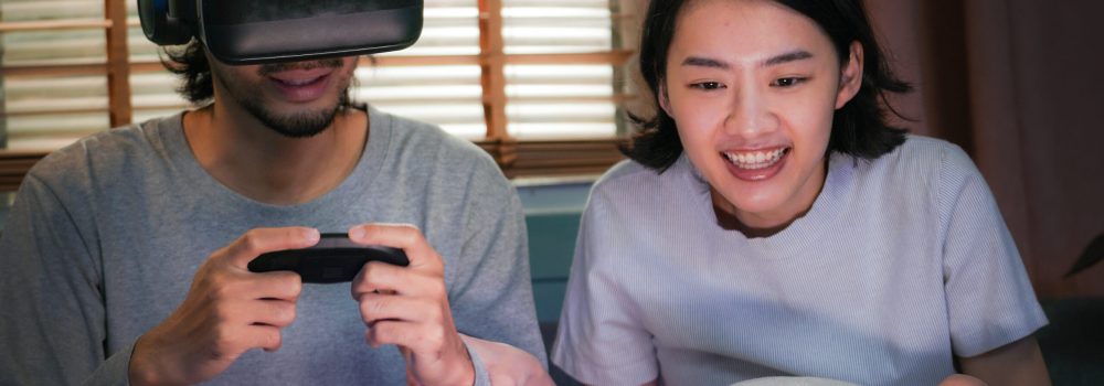 Young happy asian couple playing video games virtual reality glasses in living room. Cheerful people having fun with new trends technology. Man with hands up wearing the virtual reality goggles.